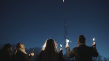 men and women holding sparklers and fireworks burst in the nights sky 