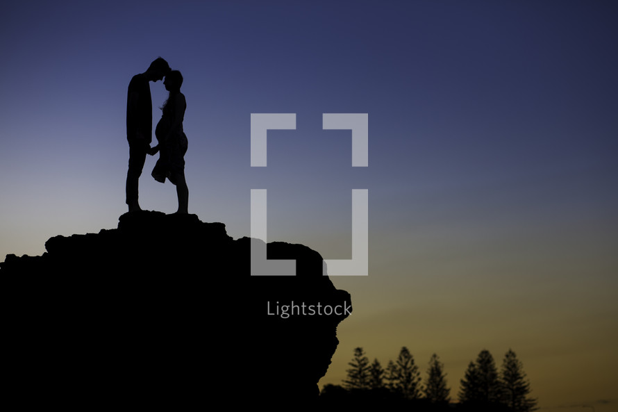 silhouette of an expecting couple standing at the edge of a cliff at dusk 