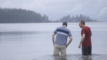 A man leads another younger man into water to be baptized in the sea.