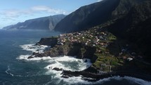 Aerial drone view village in coast of Madeira Portugal, waves crashing on a rocky shore with dramatic sea cliffs in the background