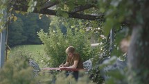 A pan through greenery outside with a man sitting and reading Bible under grape vines 