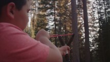 Young boy shooting a slingshot in the forest. - 1 of 2