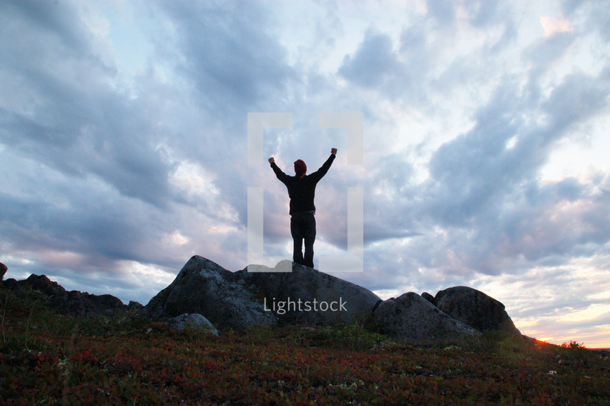 Silhouette of a man with arms raised in praise, standing on a rock outside under the clouds.
