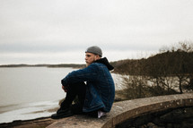 a man sitting on a wall looking out at a lake under a gloomy sky 