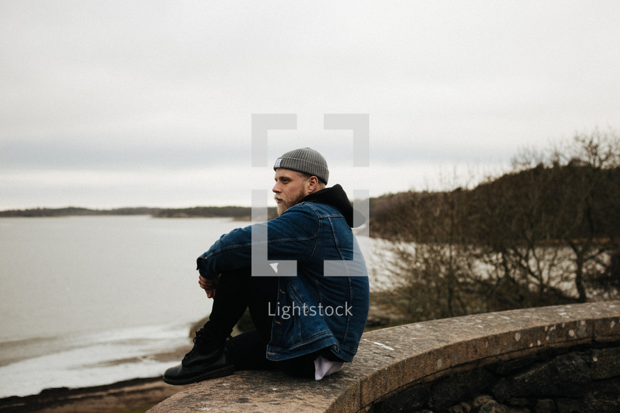 a man sitting on a wall looking out at a lake under a gloomy sky 