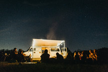 lights on a camper and friends gathered around a campfire 
