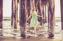 A woman walking in the water under a pier at a beach.