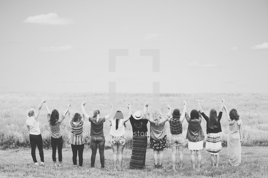 Women holding hands with arms raised in praise while standing in a field.
