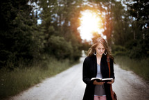 a woman standing in the middle of a dirt road reading a Bible 
