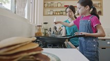 Two young girls preparing pancakes in the kitchen using a frying pan