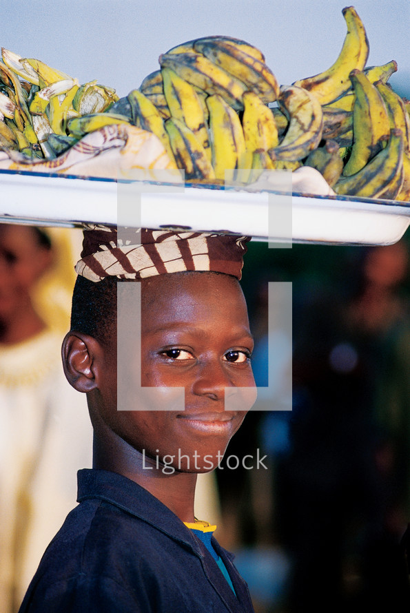 A smiling boy carrying a plate of bananas on his head.