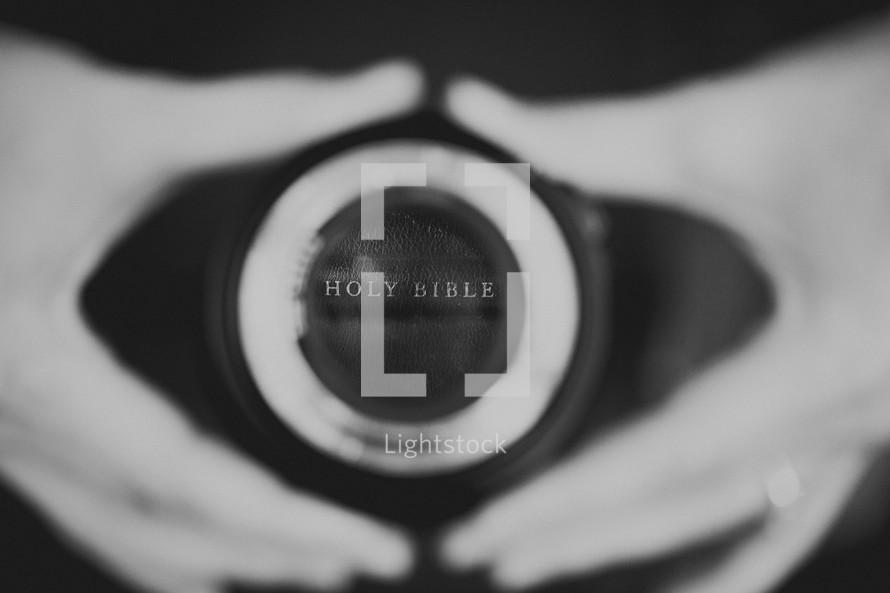 Hands holding a camera lens reflecting Holy Bible.