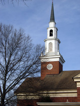 A church with a large steeple and bell tower on top of a brick church located in Virginia during a winter day against a blue clear sky and trees. 