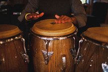 Hands beating on bongo drums in dimly-lit room.