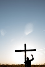 man standing in a field next to a cross with hand raised 