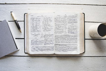 pages of a Bible, pen, journal, and coffee cup on a white wood background 