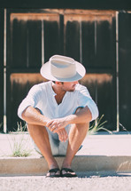 a man sitting on the curb in a cowboy hat and flip flops 