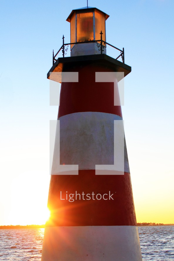 A  lighthouse with red and white stripes stands over an inlet of water and land as the sun sets behind it casting a ray of yellow and gold light against the sky and horizon.  