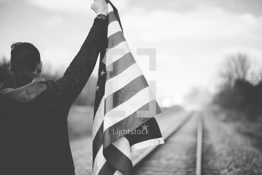 man standing on train tracks holding an American flag 