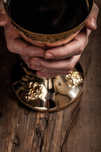 Hands holding a golden goblet of wine on a wooden surface.