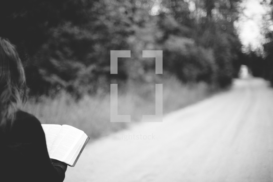 a woman reading a Bible standing in the middle of a dirt road 