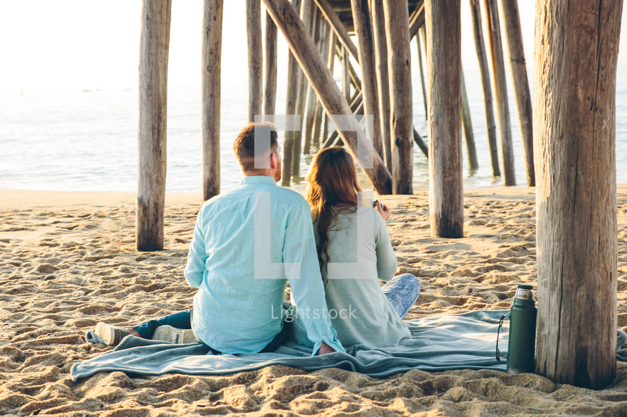 a couple sitting on a blanket under a pier on a beach 