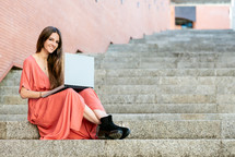 young woman sitting on outdoors stairs wearing red dress with a laptop 