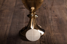 A golden wine goblet and a communion wafer on a wooden surface.