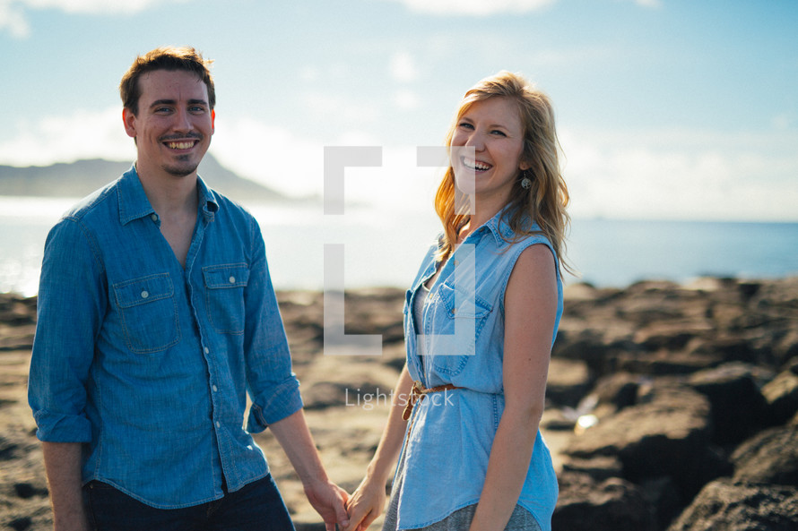 Smiling couple holding hands while standing on the ocean shore outside.