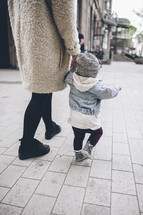 mother and toddler daughter walking holding hands on a sidewalk 