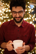 a man holding a mug of hot cocoa standing in front of a Christmas tree 