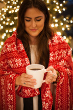 a woman wrapped in a blanket holding a mug in front of a Christmas tree 
