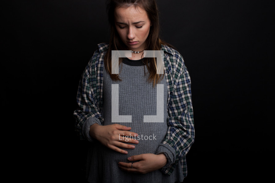 A pregnant teenager looks at her stomach with a solemn look.