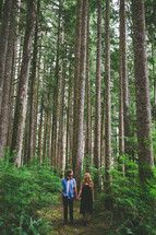 A man and woman walk hand-in-hand through a forest of tall trees.