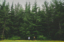 man and a woman standing in a meadow in front of a forest 