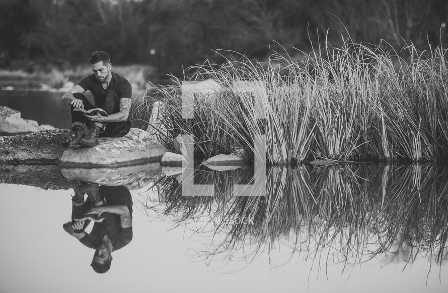 A man sitting at the shore of a pond reading a Bible outdoors 