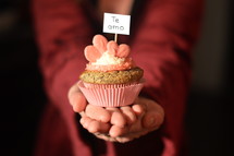 Woman offering a cupcake with pink hearts and a sign saying TE AMO.