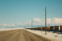 Freight train on the railroad tracks along side a dirt road with snow-covered mountains in the background.