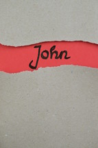 John - torn open kraft paper over light red paper with the name of the Gospel of John