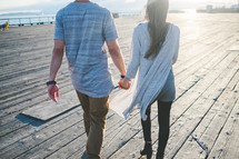 A man and woman holding hands and walking along a boardwalk.