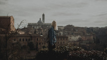 a woman with a backpack standing with an view of an ancient Eurpean City in the background 