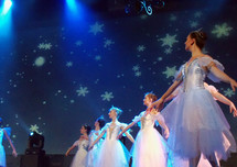 A group of female ballerina dancers from the Ukraine dance on stage wearing white outfits while surrounded by blue light and snowflake light effects on stage during a performance of The Nutcracker Christmas performance at a local church.