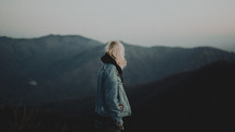 a woman in a jean jacket standing on a mountaintop 