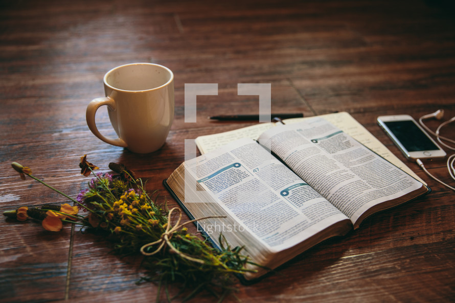 open Bible, journal, pen, coffee mug, flowers, earbuds, and iPhone on a table