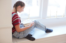 girl sitting in a window seat reading a book 
