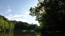 Drone footage over a small lake or pond looking up at green trees with a sunflare and a beautiful blue sky on a summer day.