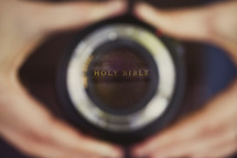 Hands holding a camera lens reflecting Holy Bible.