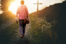 a man walking carrying a Bible on a gravel path walking towards a cross