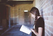 a woman leaning against a brick wall reading a Bible 