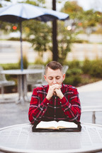 young man praying and reading a Bible sitting at a table outdoors 
